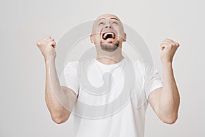 Portrait of excited upbeat bald european man with beard bending backwards while raising hands in victory gesture