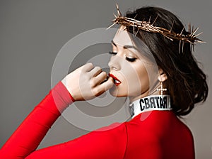 Sexy brunette woman with closed eyes and golden crown of thorns on her head in red turtleneck sweater biting her finger photo