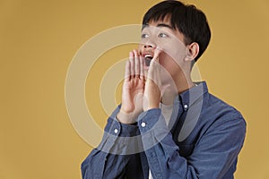Portrait of excited young asian man dressed casually with shouting announcing  holding hands near mouth isolated on yellow
