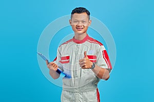 Portrait of excited young Asian mechanic holding clipboard and showing thumb up gesture isolated on blue background