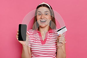 Portrait of excited young brunette woman wearing striped shirt standing with credit card and cell phone in hands, showing blank