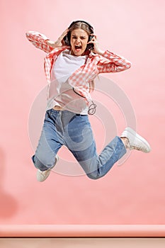 Portrait of excited young girl in checkered shirt listening to music in headphones and jumping  over pink studio