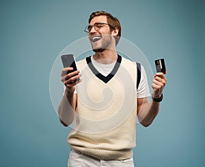 Portrait of an excited young man holding mobile phone and showing credit card isolated over blue background.