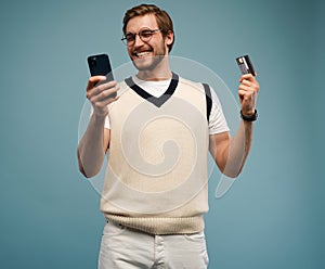 Portrait of an excited young man holding mobile phone and showing credit card isolated over blue background.