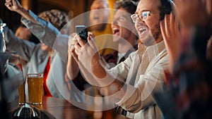 Portrait of an Excited Young Man Sitting at Bar Counter, Drinking Beer and Using a Smartphone