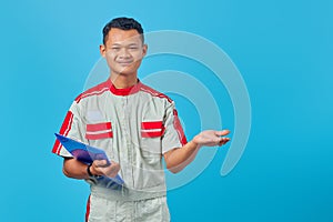 Portrait of excited young mechanic holding clipboard and pointing with palms pointing sideways isolated on blue background