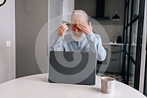 Portrait of exhausted bearded senior older businessman with grey hair touching forehead, feeling unwell due to computer