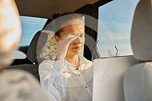 Portrait of exhausted business woman working laptop computer in car, female in white shirt using notebook in vehicle, typing on