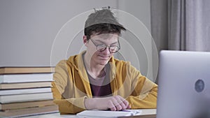 Portrait of exhausted caucasian boy in eyeglasses looking at stack of books and laptop on the table and signing. Tired