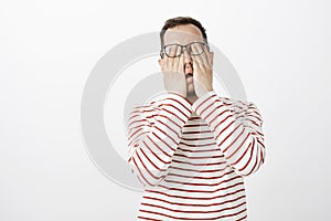 Portrait of exhausted uncomfortable male model in striped pullover and glasses, rubbing eyes, feeling pain or being