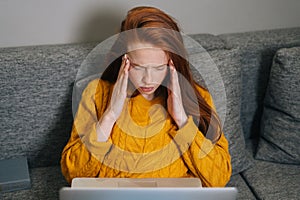 Portrait of exhausted young woman having headache while working typing on laptop computer sitting in sofa couch at home.