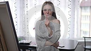 Portrait of exhausted young woman with painting brush looking at camera standing indoors at easel. Medium shot front