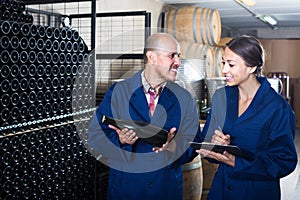 portrait of expert woman standing with clipboard and talking to winemaker in cellar