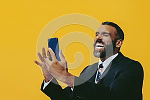 portrait of expressive angry excited bearded man in suit and tie with smartphone video call hand up yellow background