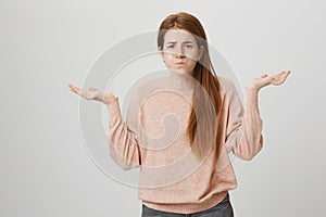 Portrait of expressive young redhead girl shrugging while being in bad mood, frowning and sulking with spreaded hands