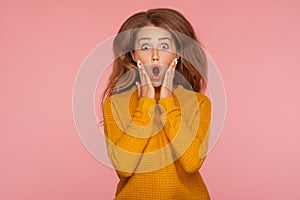Portrait of extremely excited shocked ginger girl in sweater jumping and holding hands on cheeks and screaming in amazement