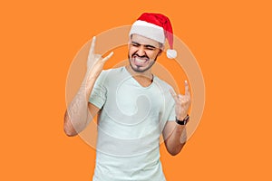 Portrait of extremely happy bearded young man in santa hat and casual white t-shirt standing with closed eyes, showing rock and