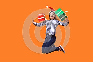 Portrait of extremely happy brunette woman in santa hat and checkered shirt jumping for joy, flying with wrapped xmas gift boxes,
