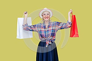 Happy cheerful mature woman in shirt, hat and eyeglasses, holding paper bags, enjoying shopping.