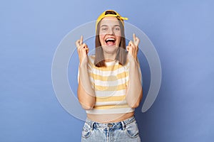 Portrait of extremely happy hopeful teenager girl wearing striped T-shirt and baseball cap standing isolated over blue background