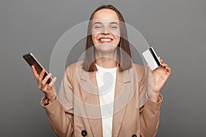 Portrait of extremely happy woman with brown hair wearing beige jacket, using cell phone and showing credit card, online shopping