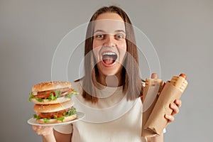 Portrait of extremely happy woman eating fast food, holding hot dog and sandwich wearing white T-shirt posing isolated over gray