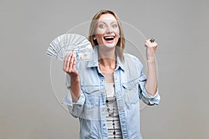 Portrait of extremely happy woman holding fan of dollars showing yes i did it gesture. isolated on gray background