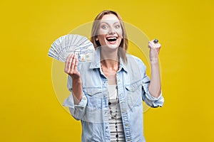 Portrait of extremely happy woman holding fan of dollars showing yes i did it gesture.  on yellow background