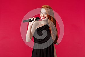 Portrait of fabulous young redhead woman with red lipstick, wear elegant evening black dress, hold microphone near lips
