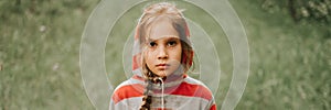 Portrait face of upset or focused thoughtful scowl eight year old kid girl in hood of striped hoodie in the forest or woodland nat