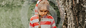 Portrait face of upset or focused thoughtful scowl eight year old kid girl in hood of striped hoodie in the forest or woodland nat