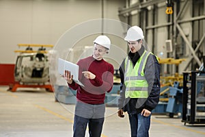 Portrait of a factory manager in a white hard hat and red sweater with laptop and factory engineer in a working clothes
