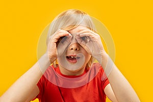 Portrait of fair-haired boy in red T-shirt keeping hands like binocular on yellow background