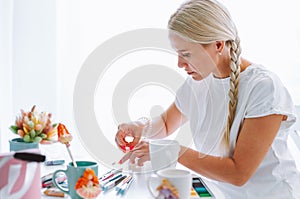 Portrait of fair-haired woman giving master class of polymer clay, jobbing and making decoration crafts flowers on mugs