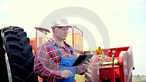 Portrait of farmer or agronomist, in red plaid shirt, is typing in tablet smth, against background of tractors