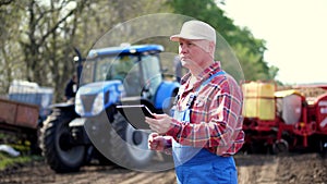 Portrait of farmer or agronomist, in red plaid shirt, is typing in tablet smth, against background of tractors