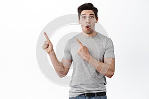 Portrait of fascinated and amazed handsome man pointing left, showing copy space for logo or banner, standing over white