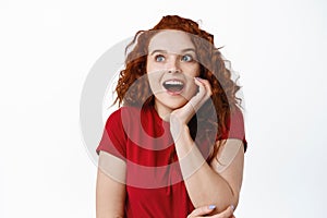 Portrait of fascinated young woman with red curly hiar, gasping amazed, staring excited at left side with copy space for