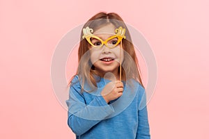 Portrait of fascinating cute little girl looking at camera through paper glasses and smiling, child playing, having fun