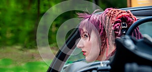Portrait of a fascinating extraordinary extravagant punk woman with pink, violet, red, dreadlocks hair in a classic sports car