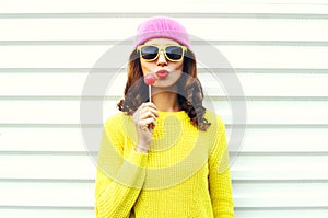 Portrait fashion pretty cool girl with lollipop blowing red lips in colorful clothes over white background wearing a pink hat