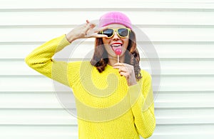 Portrait of fashion pretty cool girl with lollipop in colorful clothes over white background wearing a pink hat yellow sunglasses