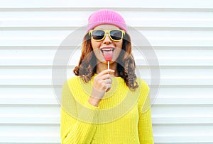 Portrait fashion pretty cool girl with lollipop in colorful clothes over white background wearing a pink hat yellow sunglasses