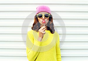 Portrait fashion pretty cool girl sucking lollipop in colorful clothes over white background wearing a pink hat yellow sunglasses