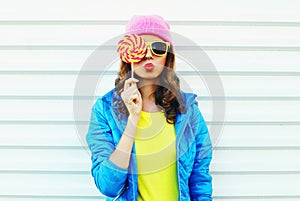 Portrait fashion pretty cool woman with lollipop in colorful clothes over white background wearing a pink hat yellow sunglasses