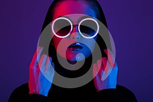 Portrait of fashion young girl in a black sweater with a hood and round sunglasses in red and blue neon light in the