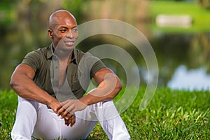 Portrait fashionable African American man sitting on grass in the park. Man is smiling and glancing of camera