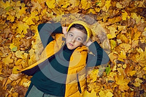 portrait of a fashionable baby boy in a yellow vest and hat in autumn lying in orange leaves in the afternoon on the