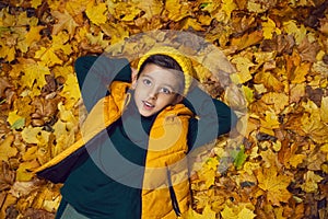 portrait of a fashionable baby boy in a yellow vest and hat in autumn lying in orange leaves in the afternoon on the