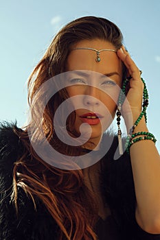 Portrait of fashionable girl in black dress and stilish tiara with green jewellry in her hand in sun light on blue sky background